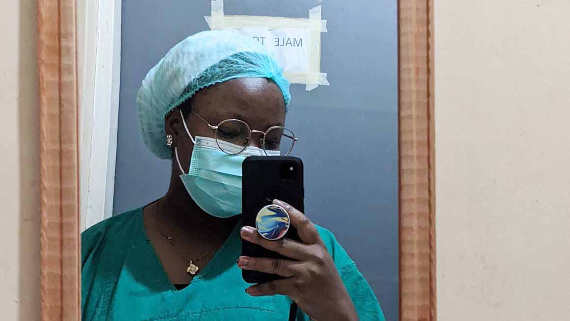A Black woman pictured in a mirro photographing herself with a phone, wearing a surgical mask and in medical clothing