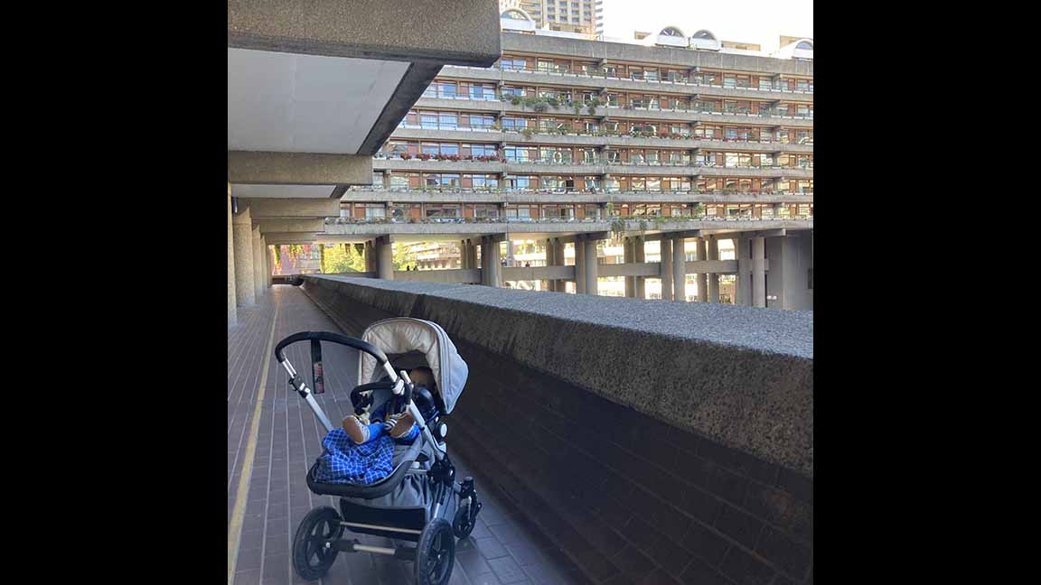 A baby in a stroller on a coomon exterior balcony of an apartment complex with many apartments visible in the background