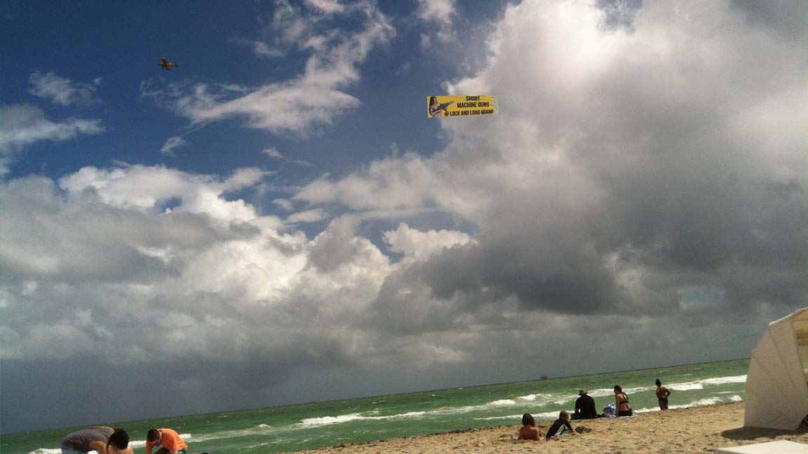 A photo of an airplane pulling a banner that reads SHOOT MACHINE GUNS @ LOCK AND LOAD MIAMI above the water as view from the beach.
