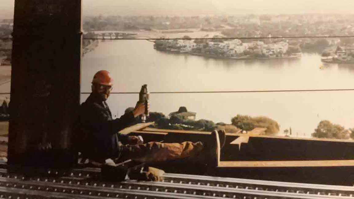 Photo of the author wearing a construction hard hat, sitting on a steel beam high up on a building under construction, smiling and holding a thermos as if they are toasting toward the camera.