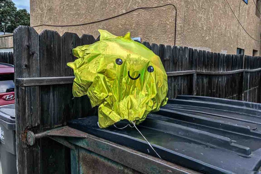 An iridescent-metallic yellow-green balloon with a smiley face floating out of a dumpster