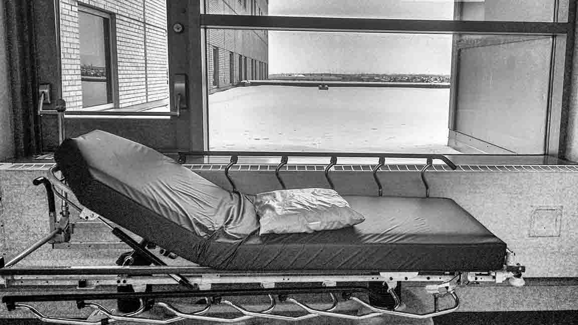 A black & white image of a hospital bed near a large window. Very gray and serious image